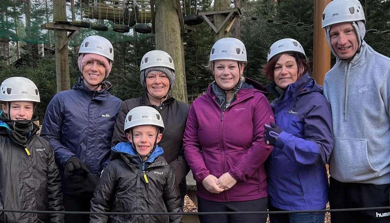 Group of seven people pose, smiling, wearing white helmets and outdoor jackets; context is a forest adventure ropes course with platforms and nets behind. Text: “PETZL”; “gelert”; “Dare2b”.