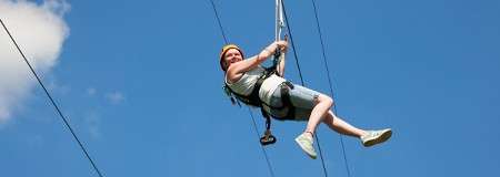 Person in a helmet and harness glides on a zipline, gripping the trolley, legs extended, against a clear blue sky with a small cloud.