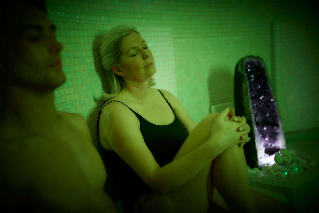 Woman in black swimsuit sits with eyes closed, relaxing against a tiled wall; a shirtless man beside her rests; purple geode and white stones glow in a green-lit steam room.