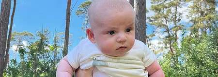 Baby in a white onesie is held up by an adult hand, gazing ahead, surrounded by tall pine trees, green foliage, and blue sky with scattered clouds.