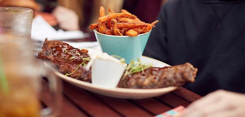Barbecued ribs rest on a plate, glistening with sauce, accompanied by a bowl of fries and a dip; set on an outdoor wooden table beside a diner, background softly blurred.
