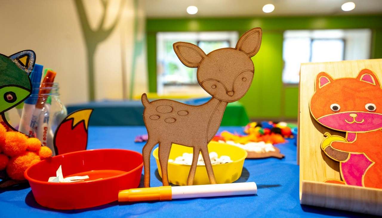 Wooden deer cutout stands upright, surrounded by craft supplies—markers, red and yellow bowls, pom-poms, and animal decorations—on a blue-covered children’s table in a brightly lit classroom.