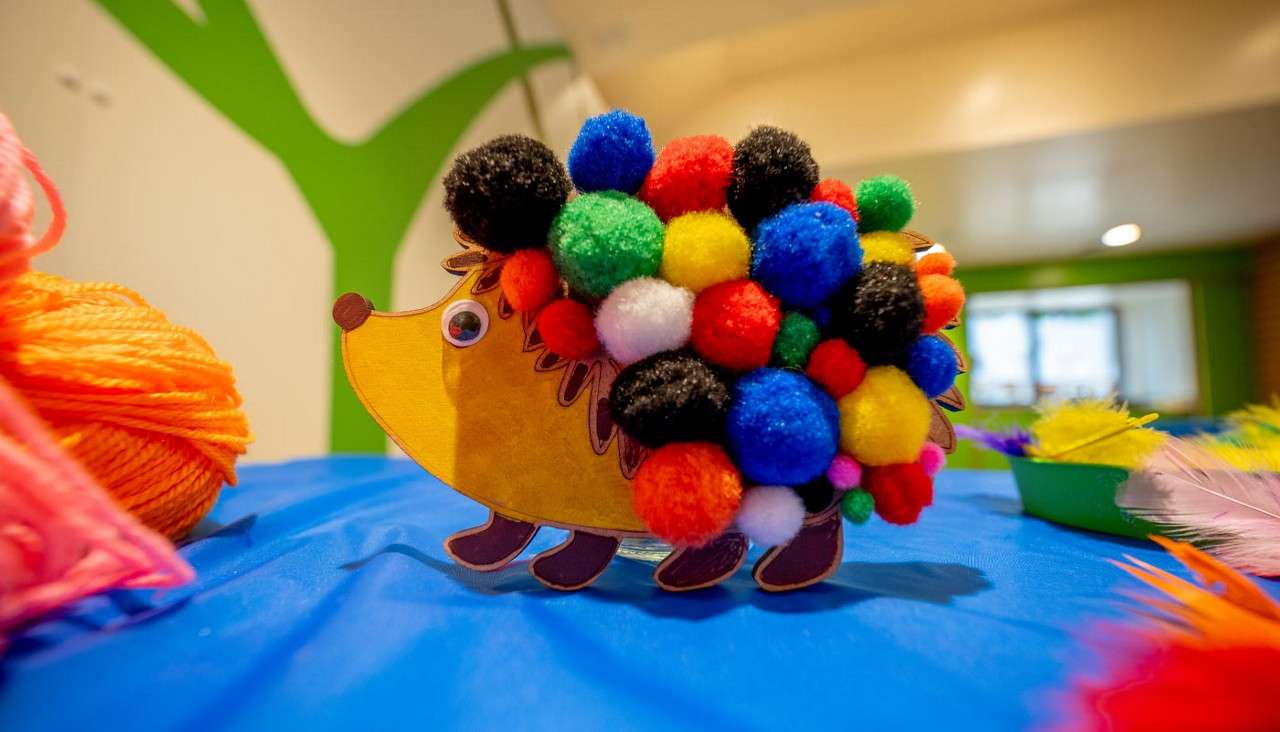 Toy hedgehog stands decorated with multicolored pom‑pom spines, displayed on a blue-covered craft table; surrounding supplies include orange yarn and feathers in a brightly lit room with green walls.