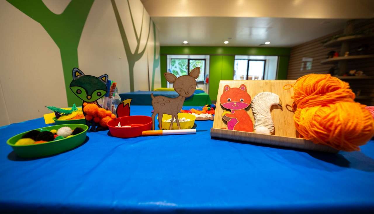 Craft supplies rest on a table; animal cutouts stand among bowls of pom‑poms, markers, and orange yarn in a brightly lit classroom with green walls and large windows.