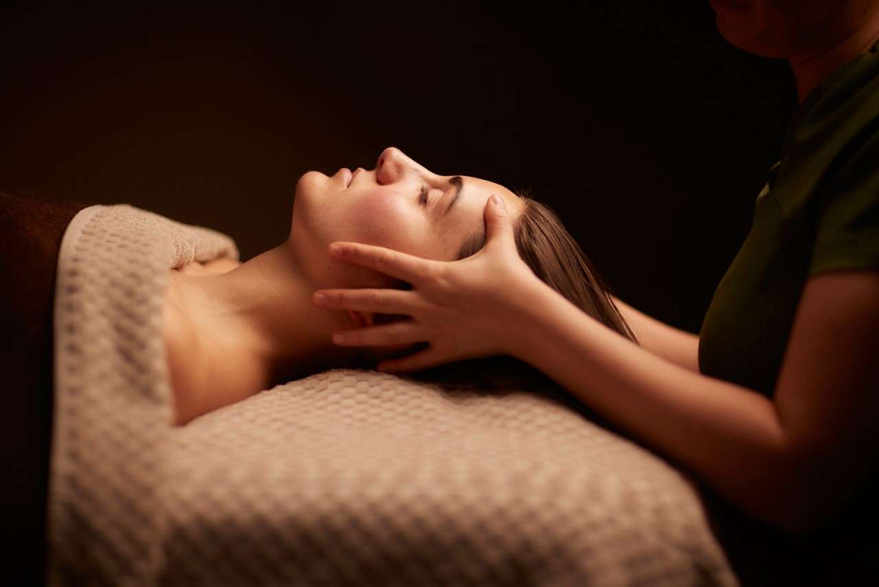 A person’s face, eyes closed, receives a gentle head massage; a therapist’s hands cradle the jaw and scalp while they lie under a textured blanket in a dim spa setting.