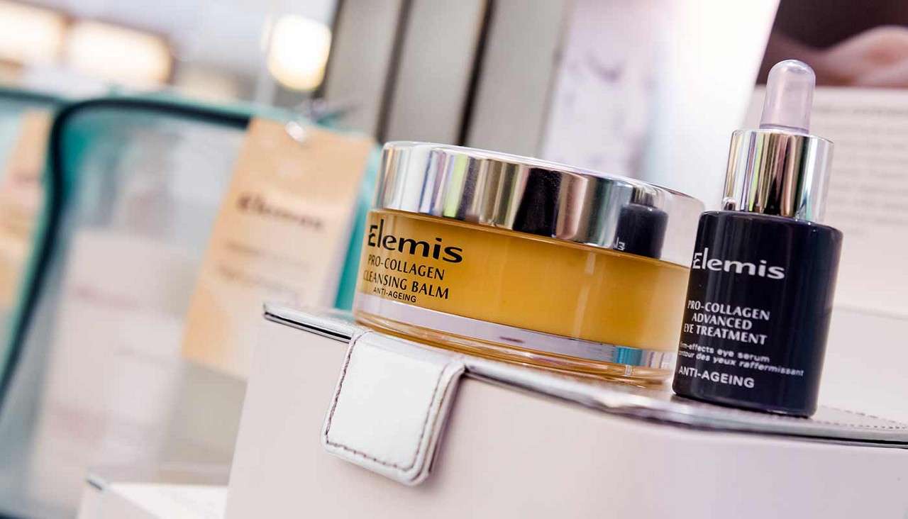 Two skincare containers rest on a white display in a shop: a yellow jar with a silver lid and a dark dropper bottle; background packaging blurred. Text: Elemis. PRO-COLLAGEN. CLEANSING BALM. ANTI-AGEING. Elemis. PRO-COLLAGEN. ADVANCED EYE TREATMENT. multi-effects eye serum. ANTI-AGEING.