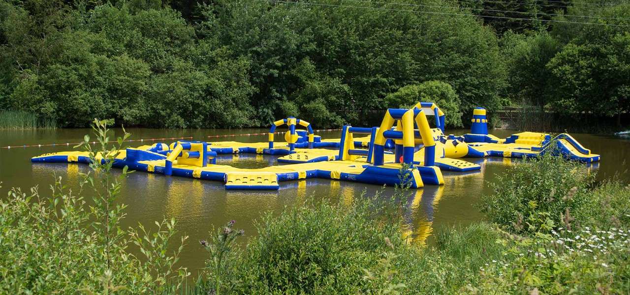 Inflatable water obstacle course floats, interconnected slides, platforms, and arches forming a circuit, in a calm pond; surrounded by dense green trees and shrubs, marked by buoy lines.