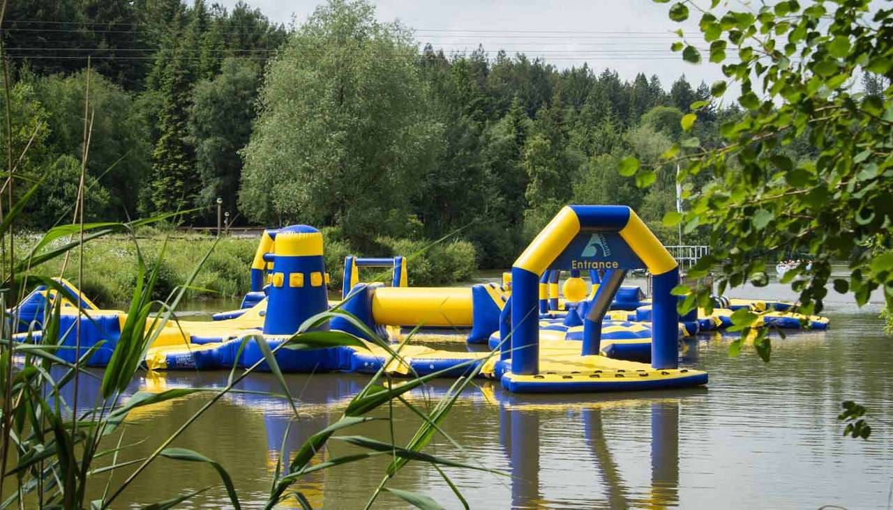 Inflatable water-park course floats, inviting play; platforms, slides, and towers connect under an arch labeled "Entrance". Context: calm lake bordered by reeds and dense trees, with distant shore structures.