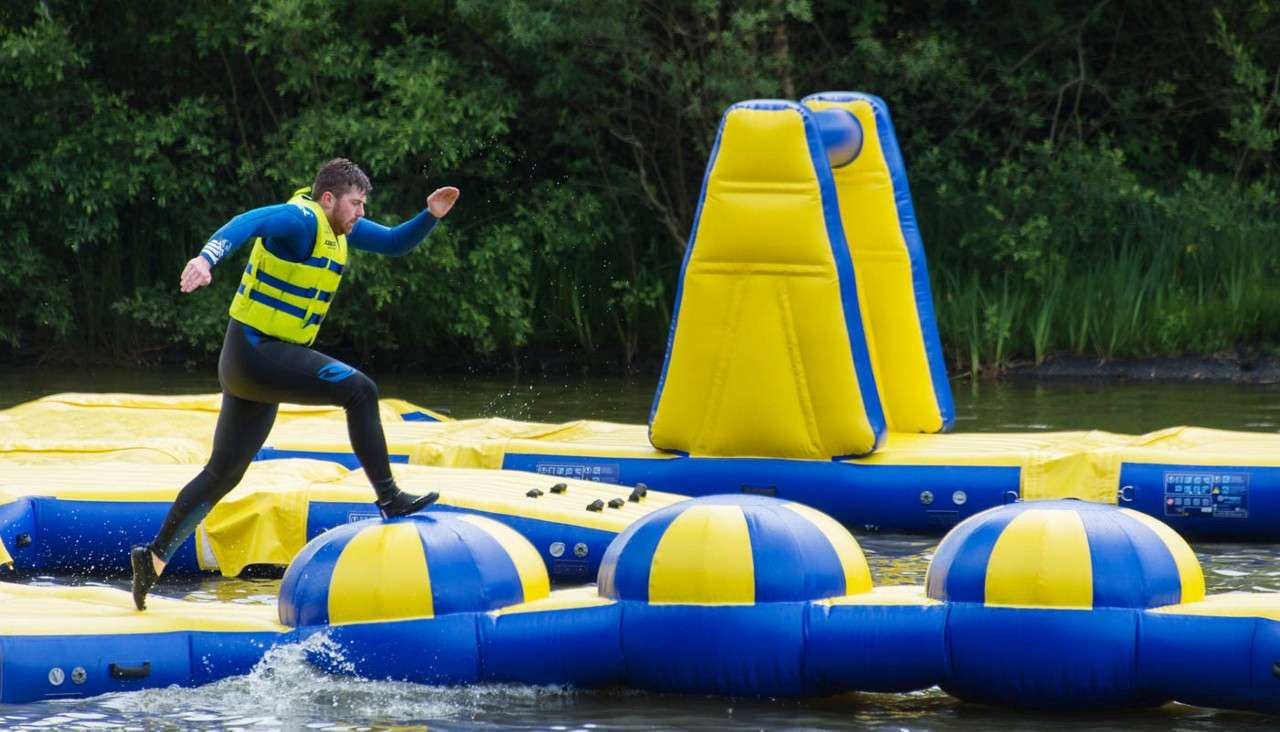 Man in a yellow life jacket leaps between blue-yellow inflatable domes. A floating water obstacle course spans a calm lake, with larger inflatables nearby and trees along the shore.