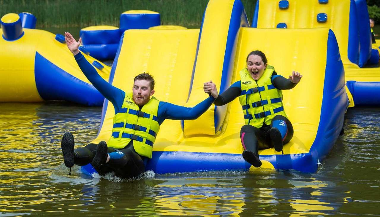 Two adults hold hands, slide feet-first into water, splashing, while wearing bright life jackets and wetsuits on a yellow-and-blue inflatable water-park obstacle. Visible text: JOBE (on both vests).