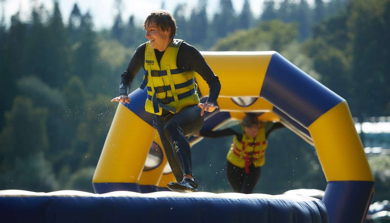 Runner in yellow life vest leaps across a wet inflatable water-course platform, another participant crawling through a blue-yellow arch behind; forested shoreline and lake in background. Visible text: JOBE.