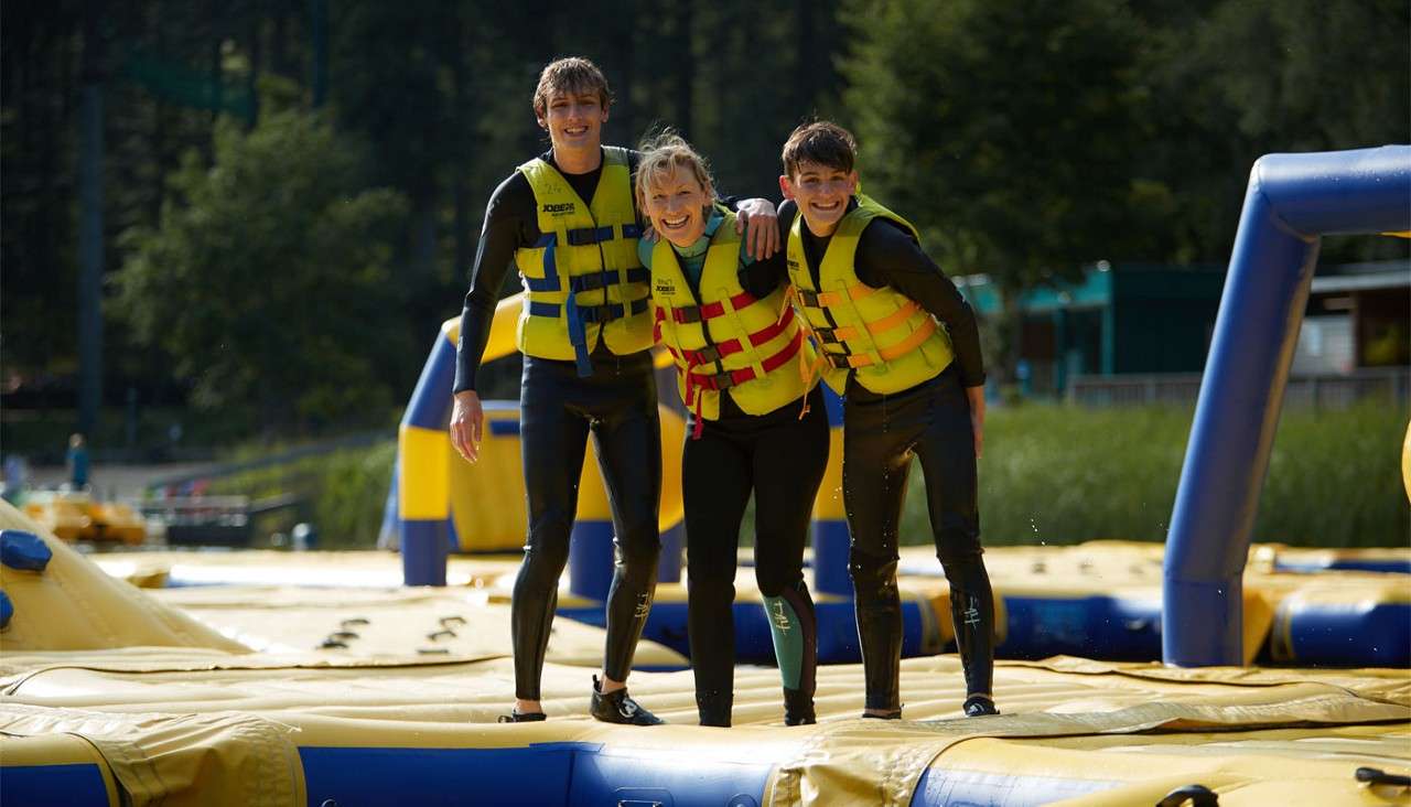 Three wetsuit-clad people stand arm-in-arm, smiling. They balance on a yellow-and-blue inflatable water park obstacle. Forested park and facilities sit in the background. Visible text: “JOBE” on life jackets.