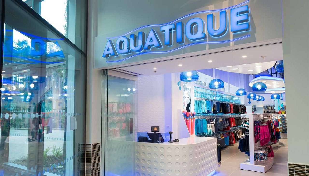 Storefront displays merchandise; racks of colorful swimwear line aisles under blue dome lights, behind a white reception counter. Context: modern glass entrance with illuminated sign reading AQUATIQUE.