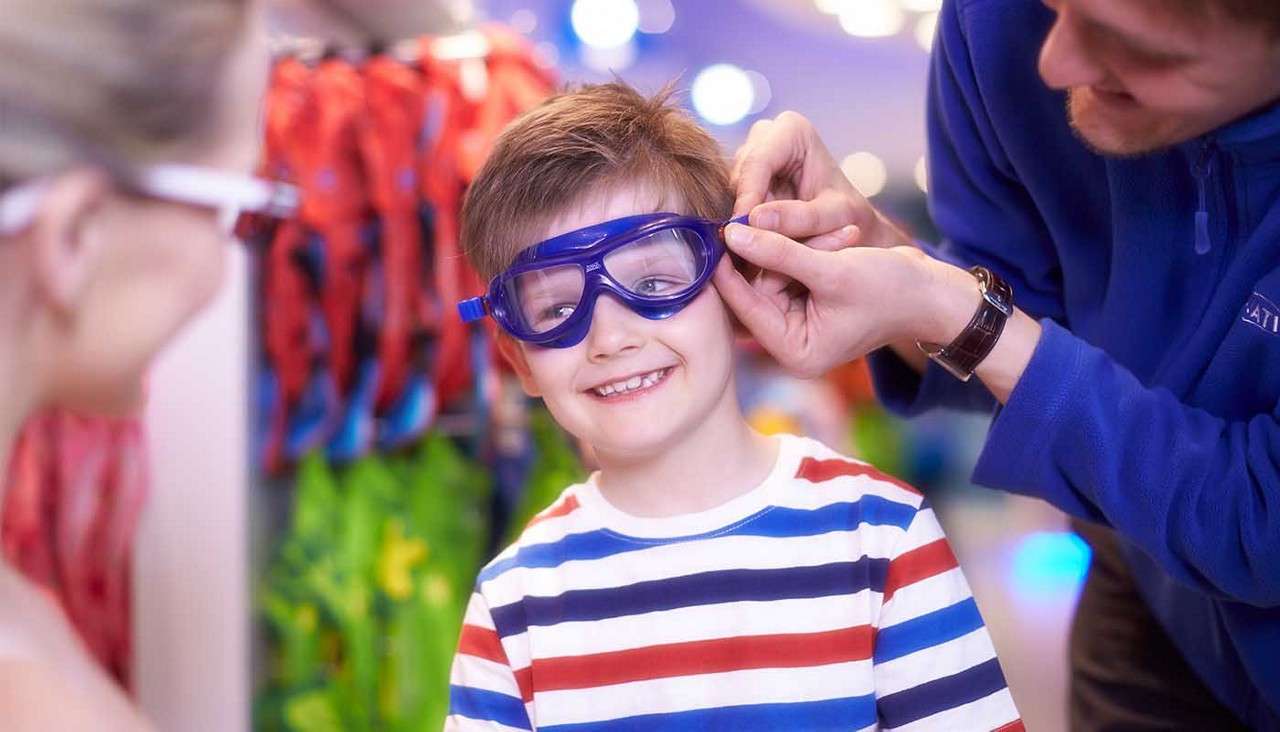 Boy wearing blue swim goggles smiles while an adult adjusts the strap, in a brightly lit store aisle with colorful swim gear hanging blurred in the background.