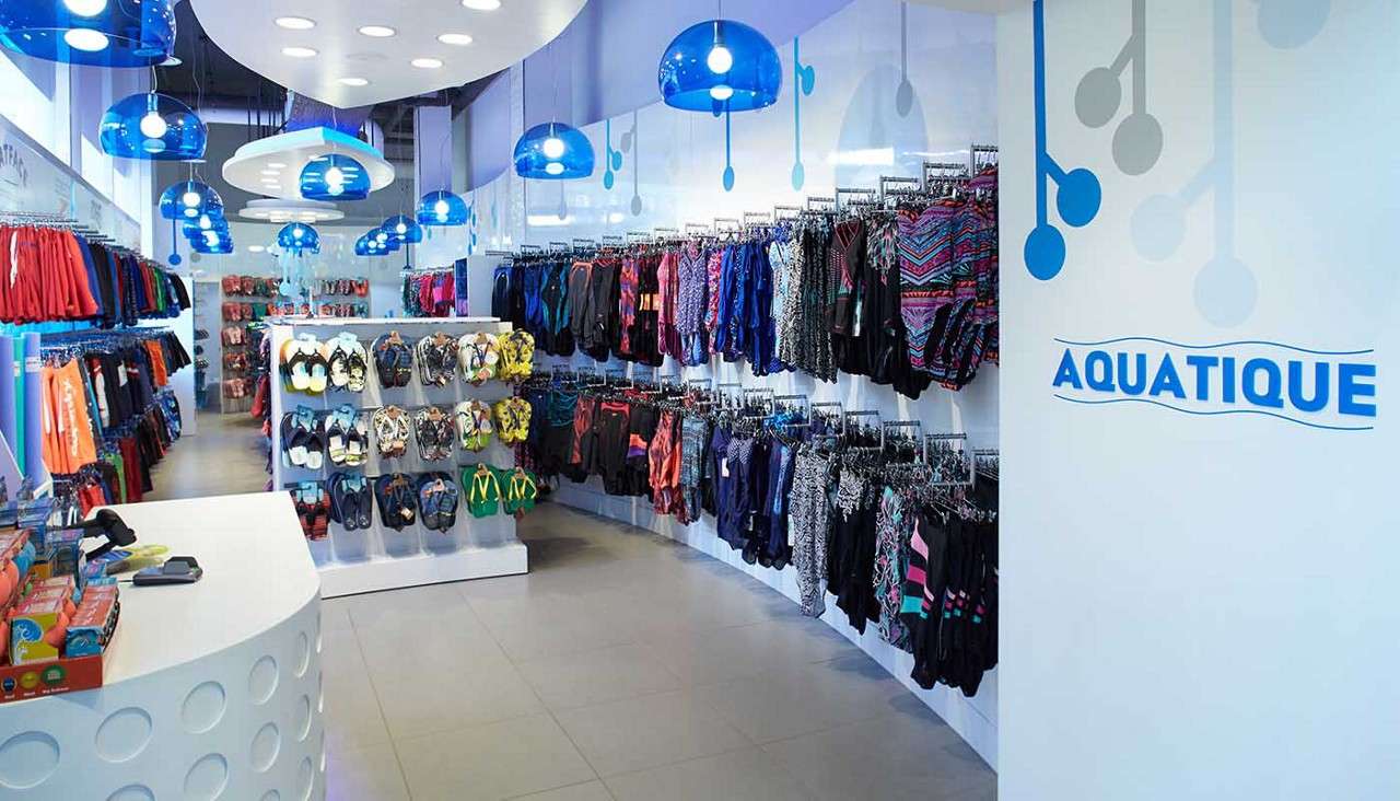 Swimwear displays hang and line the walls, showcasing colorful suits and flip-flops; bright blue pendant lights illuminate a sleek retail store. Visible text: AQUATIQUE.