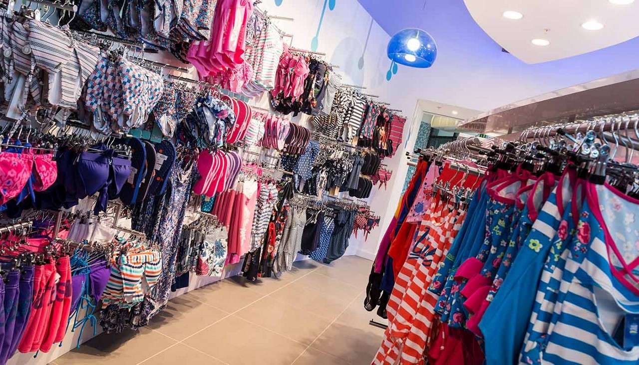 Swimwear racks display colorful bikinis and one-piece suits, hanging neatly on hangers, in a brightly lit retail store aisle with tiled floor, patterned walls, and overhead lights.