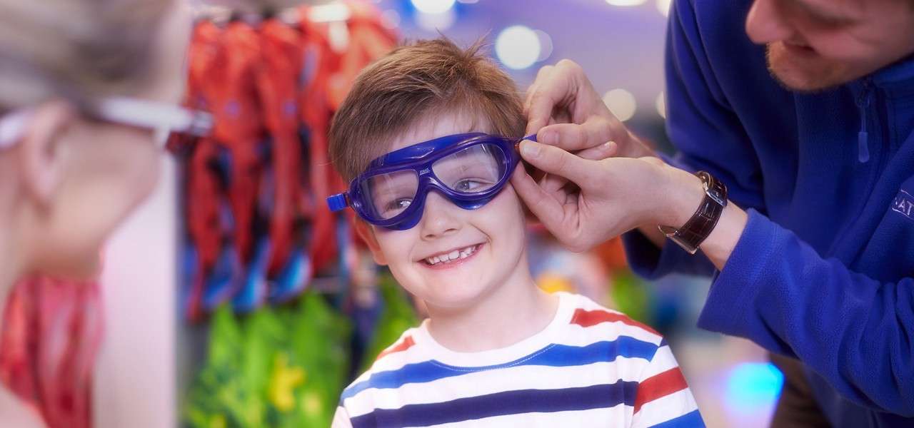 Boy wearing blue swim goggles smiles while an adult adjusts the strap; another adult watches nearby, in a brightly lit store with colorful swim gear hanging in the background.