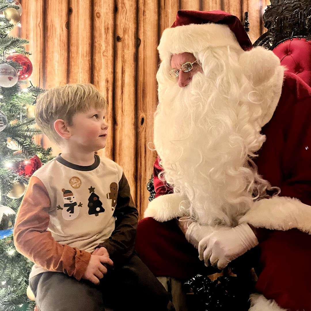 Boy sits facing a man dressed as Santa, listening as Santa leans in, gloved hands together, beside a Christmas tree with ornaments, against a wooden backdrop on a red chair.