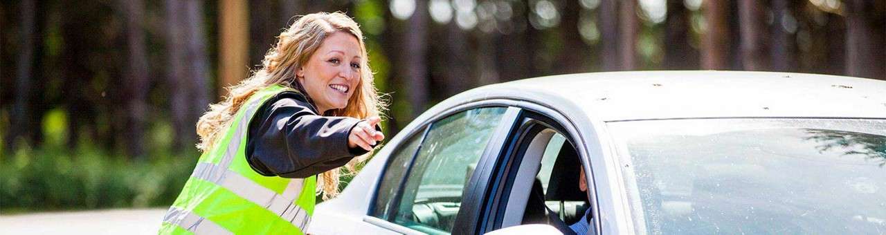 Traffic marshal in a neon reflective vest gestures and smiles toward a driver in a white car, guiding traffic on a road bordered by tall trees in daylight.