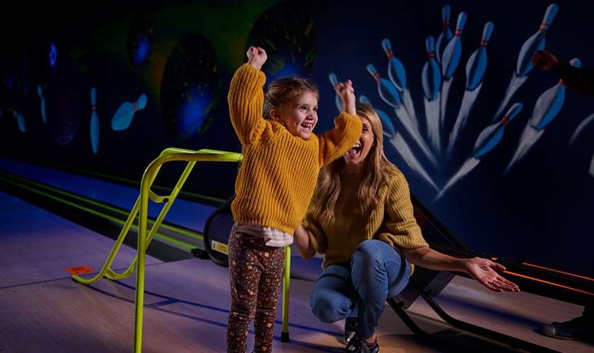 Child celebrates with arms raised, smiling. Adult beside her kneels and claps. In a dim bowling alley, a green ball ramp faces the lane, with neon pin murals in background.