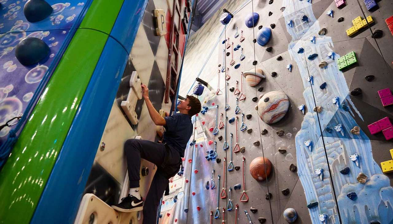 A climber ascends handholds, reaching upward; surrounding walls display cartoon planets, bubbles, and geometric grips in a busy indoor climbing gym. Visible text: P.