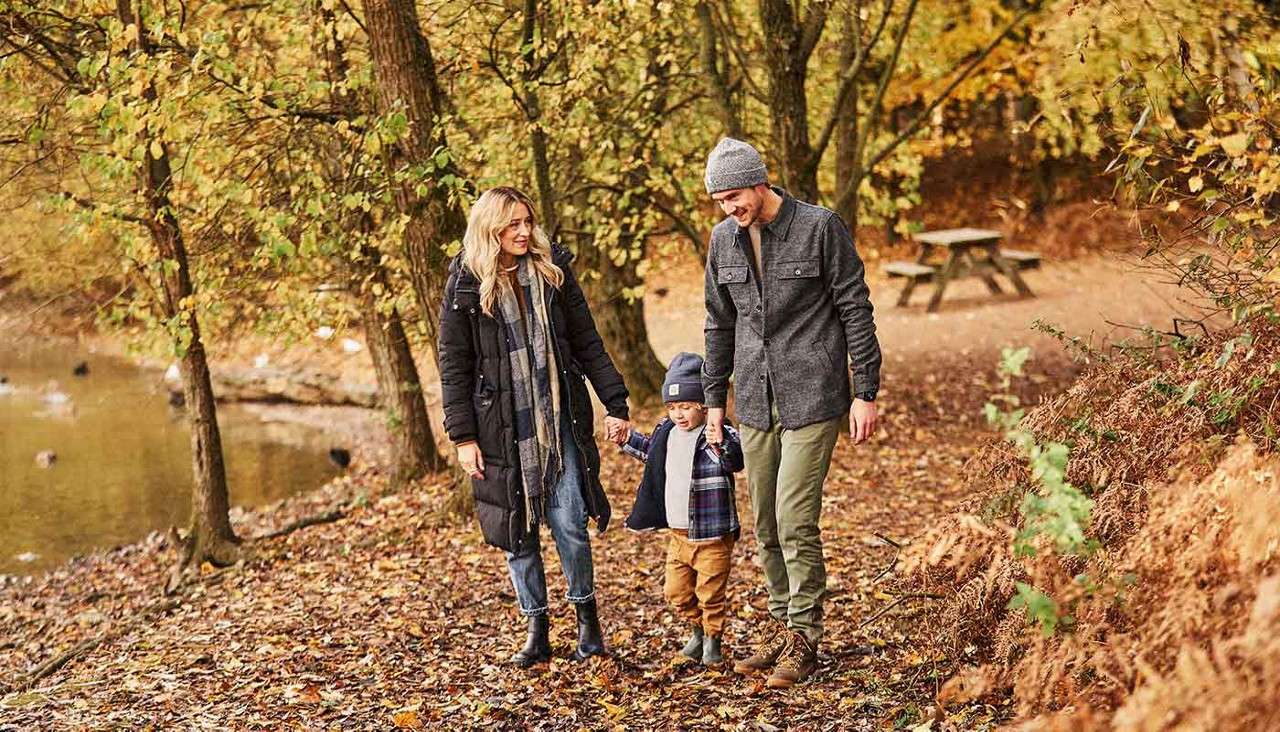 Family of three walks hand-in-hand, smiling, along a leaf-strewn woodland path beside a pond with ducks. Autumn trees surround them; a picnic table sits in the background.