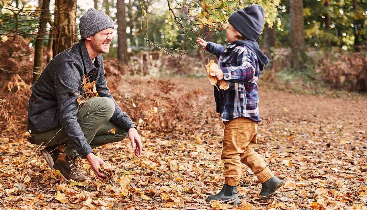Man and small child toss fallen leaves together, smiling. They wear hats and jackets. Context: woodland trail in autumn, ground blanketed with orange leaves and trees surrounding.