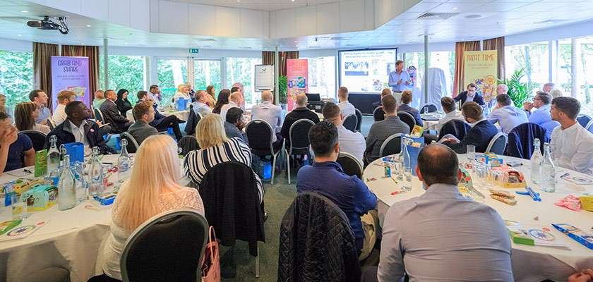 Presenter addresses a projected slide, while attendees sit at round tables listening and taking notes in a bright conference room with large windows, banners, and refreshments.