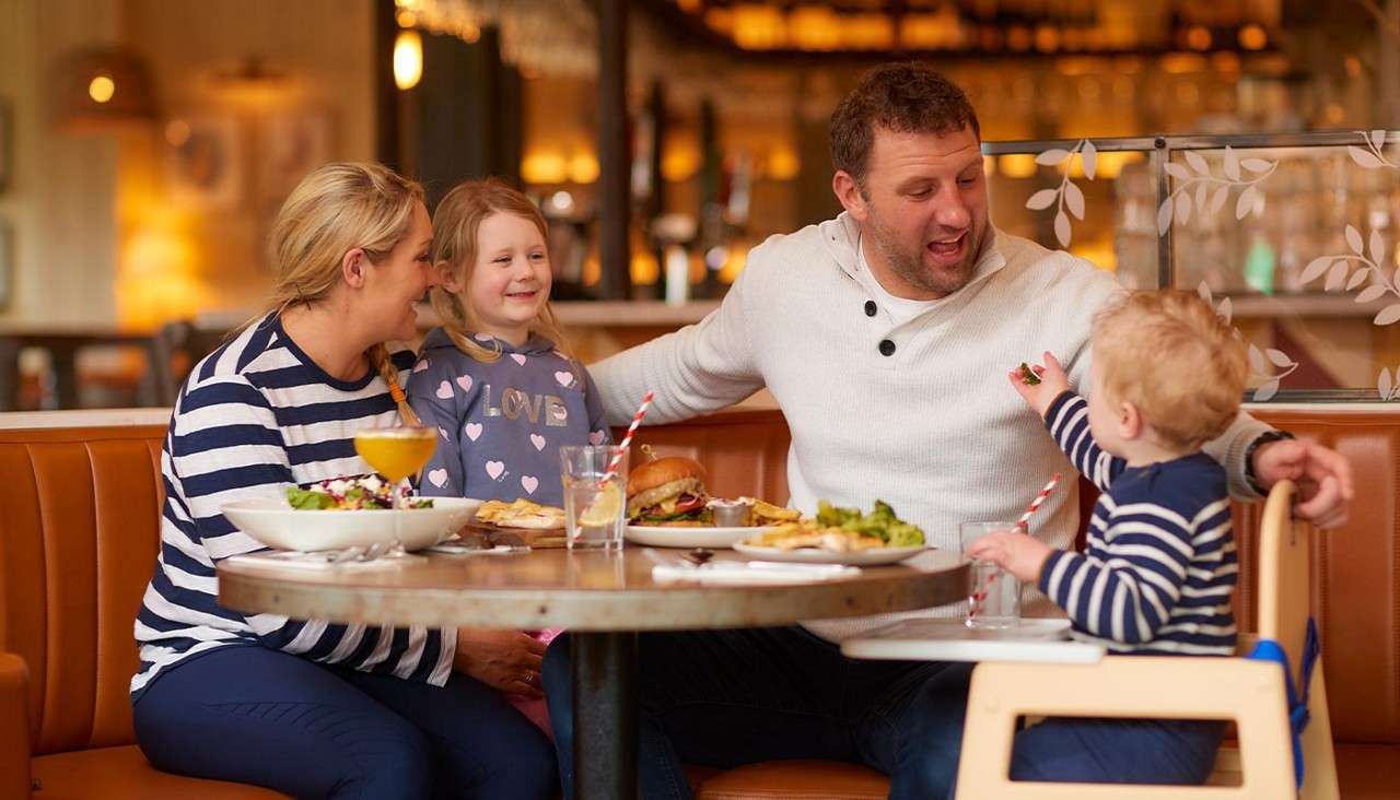 Child offers food to father at restaurant table; parents and sister watch, smiling. Warmly lit booth, plates with burger, fries, salad, drinks with striped straws. Girl’s sweatshirt text: "LOVE".