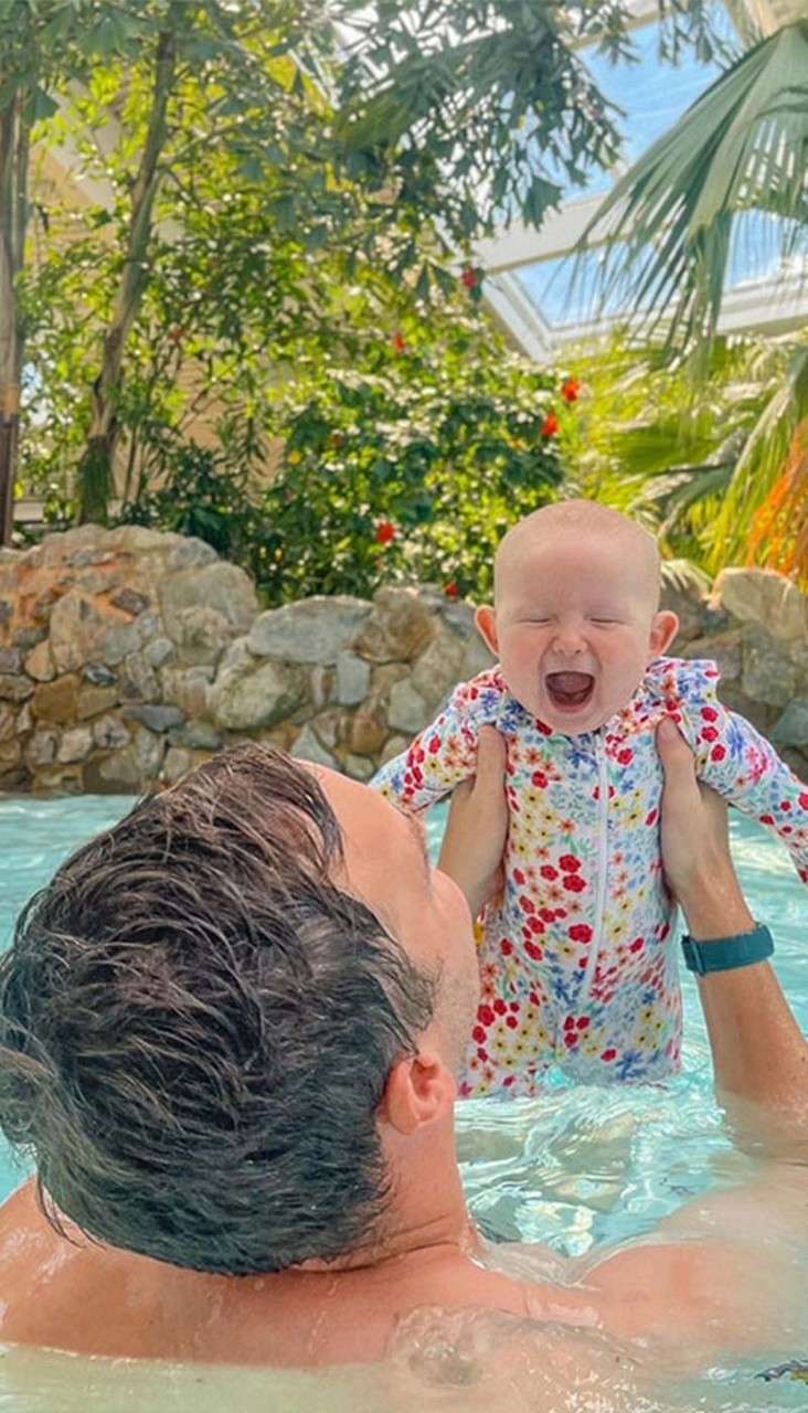 Baby in floral swimsuit laughs with mouth open while being lifted by an adult in a pool, surrounded by rocks, tropical plants, and a sunlit glass-roofed conservatory.