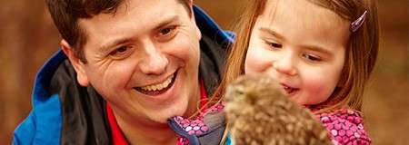 Owl perches calmly on a gloved hand; a smiling man and young girl watch closely beside it, outdoors with a warm, blurred brown background.