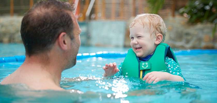 Child in a green flotation vest laughs and splashes, reaching toward an adult nearby; context: indoor swimming pool with blue water, stone wall, fencing, and plants in the background.
