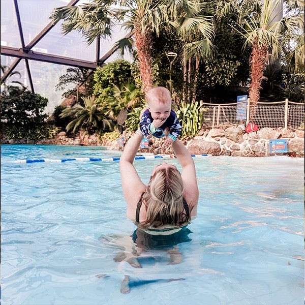 Baby is lifted above the water by an adult in a shallow swimming pool; palm trees, rocks, rope barriers, and a skylit indoor tropical enclosure surround them.
