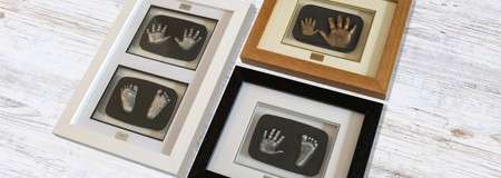 Framed baby hand and foot casts sit side-by-side; bronze-toned imprints displayed in white, black, and wood frames on a whitewashed wooden surface.