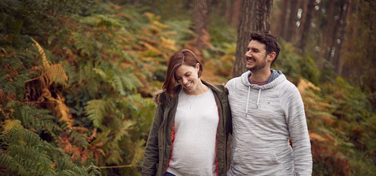 Two adults walk closely, smiling, one with an arm around the other's shoulders, in a fern-filled forest with tall trees and autumnal foliage; casual jackets suggest a cool day.