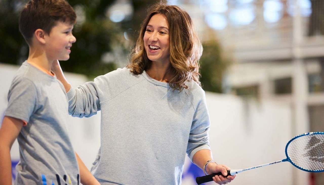 Woman encourages a boy, smiling and touching his shoulder, while holding a badminton racket; indoors on a bright gym court with blurred walls and windows.