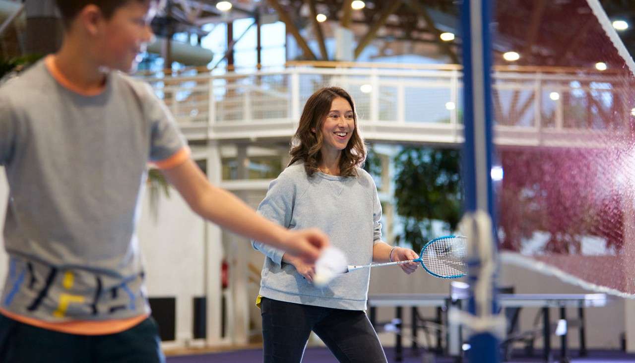 Two players with badminton rackets rally, smiling and moving near a net, in an indoor sports center with high ceiling, walkway railings, bright lights, and tables visible in the background.