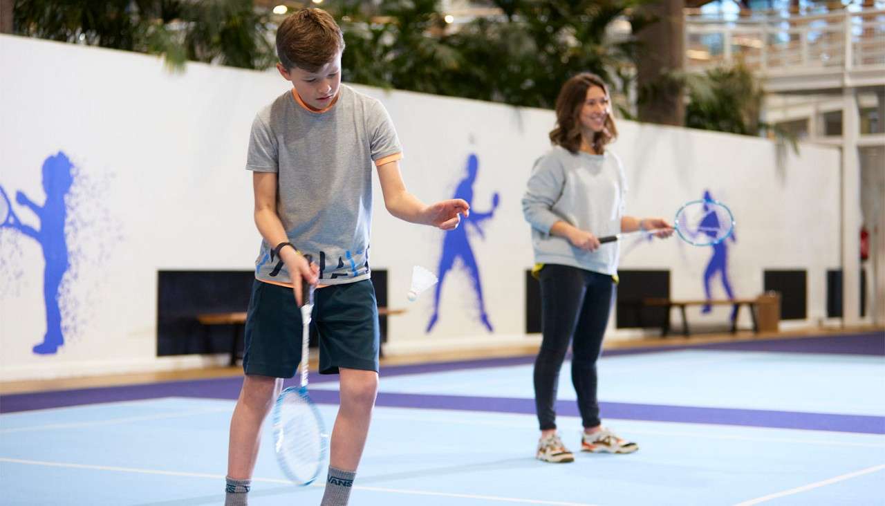 Two badminton players bounce a shuttlecock with rackets on an indoor blue court; wall silhouettes and benches behind. The nearer player wears socks labeled “VANS.”
