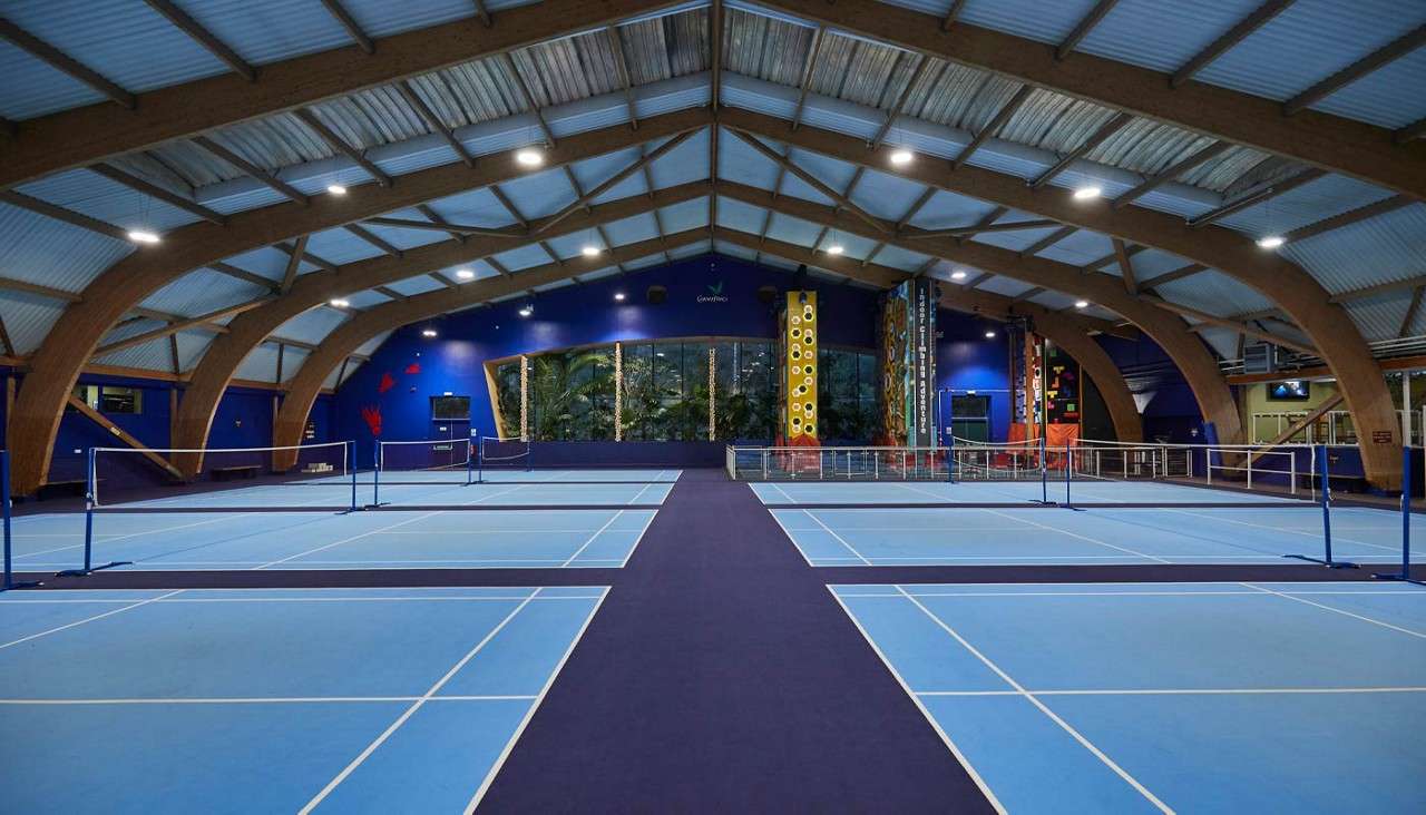 Badminton courts sit empty, nets stretched; in the background, colorful climbing walls rise inside a spacious arched-roof recreation hall. Text: “Clip ’n Climb”.