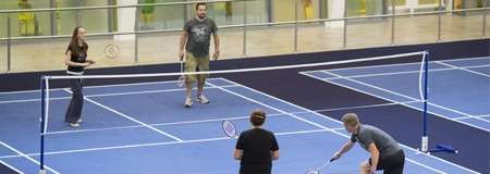 Four adults play badminton; two teammates return a shot while opponents ready near baseline. Indoors on blue courts with white lines, bordered by railing and windows, indoor track visible.