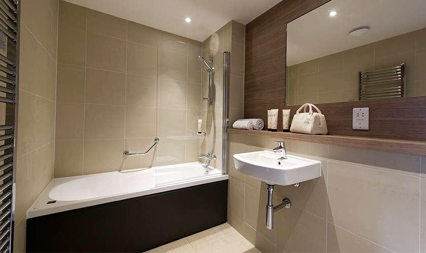 Bathtub with showerhead, glass screen, and grab bar sits beside a wall-mounted sink and mirror; beige tiles, wooden ledge, towels, toiletries, and heated towel rail surround in a modern bathroom.
