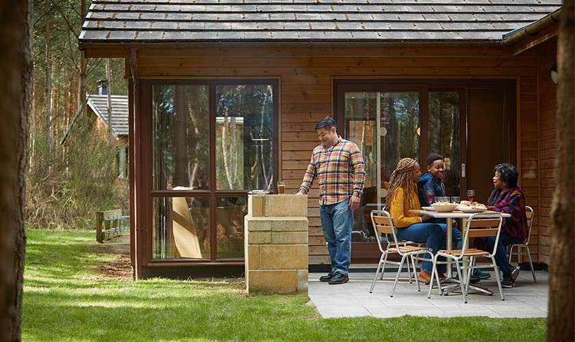 Group of four people eat at a patio table while another tends a stone grill, outside a wooden cabin, surrounded by trees and grass in daylight.