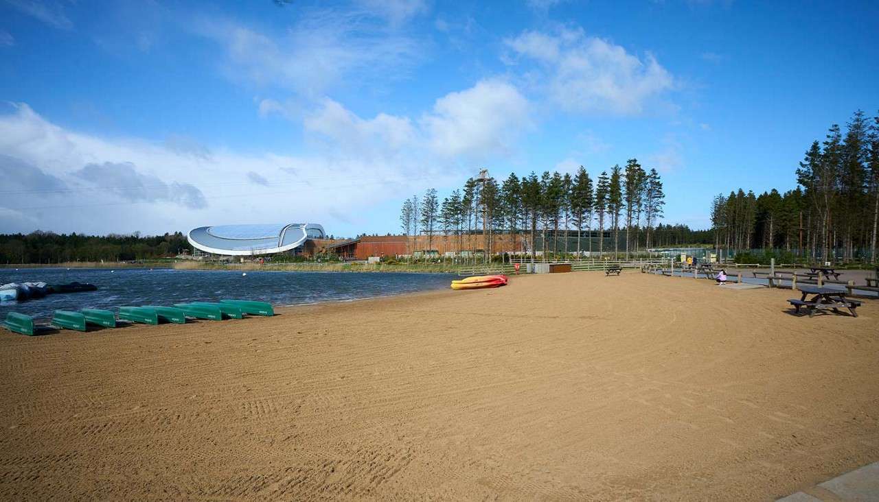 Empty sandy beach borders a small lake; canoes and kayaks rest ashore. Background holds picnic tables, tall pine trees, and a modern curved-roof building beneath a blue, partly cloudy sky.
