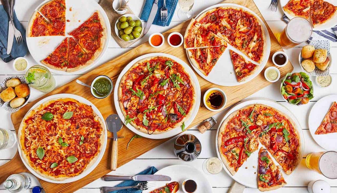 Pizzas are sliced and served on plates and a wooden board, shared alongside dips, olives, salad, bread rolls, drinks, and utensils across a bright white tabletop.