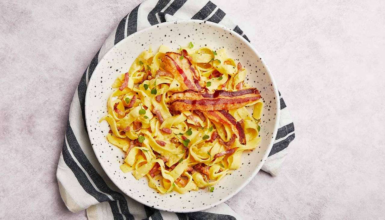 Creamy tagliatelle pasta sits topped with crisp bacon strips and scattered herbs, served in a speckled white bowl resting on a black-and-white striped cloth over a pale surface.