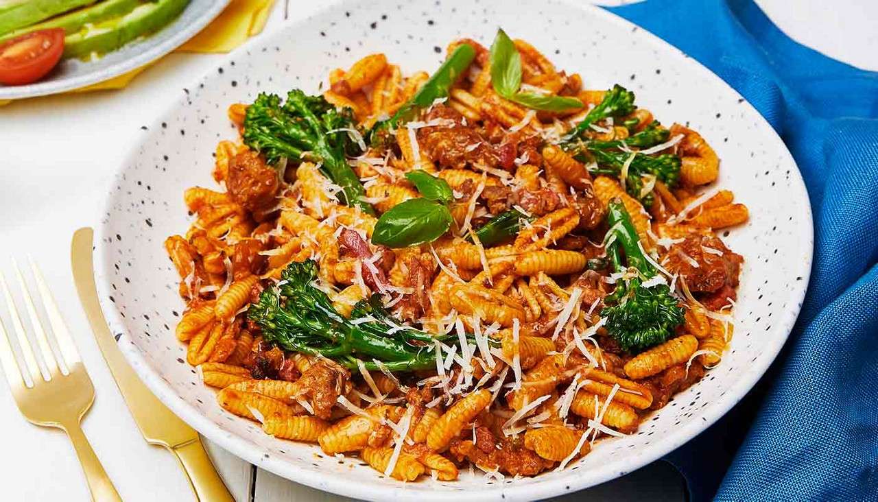 Ridged pasta tosses with beef ragu and broccolini, topped with grated cheese and basil, resting in a speckled white bowl on a table with gold cutlery and a blue napkin.