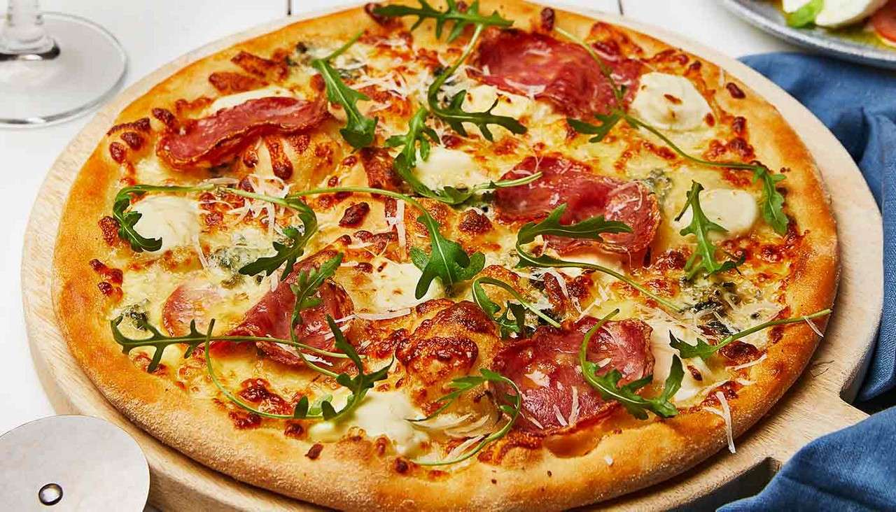 Pizza rests on a wooden board, topped with melted cheese, salami slices, arugula, and dollops of sauce; a metal pizza cutter, wineglass stem, and blue napkin sit on a tabletop.