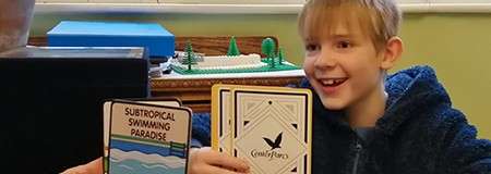 Child presents cards, smiling, in a room with a window and toy model on a table. Visible text: SUBTROPICAL SWIMMING PARADISE; Center Parcs.