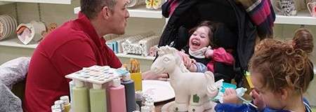 Adult and two children paint ceramics; a stroller-seated child laughs as the adult engages, in a pottery studio with shelves, paint bottles, and a ceramic unicorn centerpiece on the table.