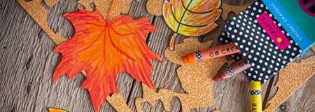 Paper leaves and glittery cutouts lie scattered; crayons in a polka-dot pouch rest nearby on a wooden table, suggesting an autumn craft activity. Visible text: GO.